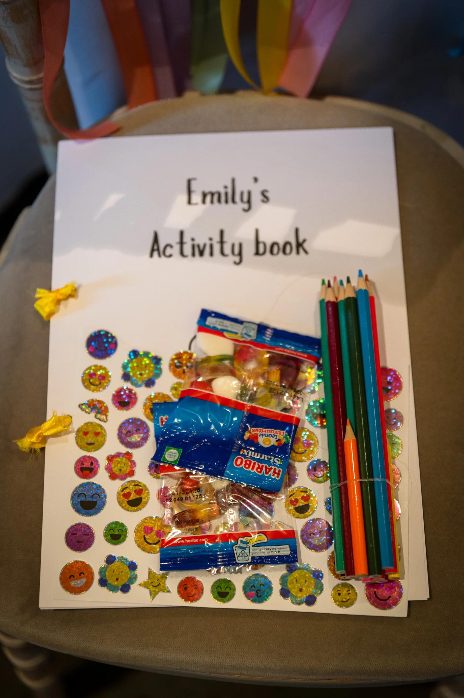 A flat lay image of a personalised children's activity book with colourful stickers, pencils, and sweets resting on a chair at a wedding reception.