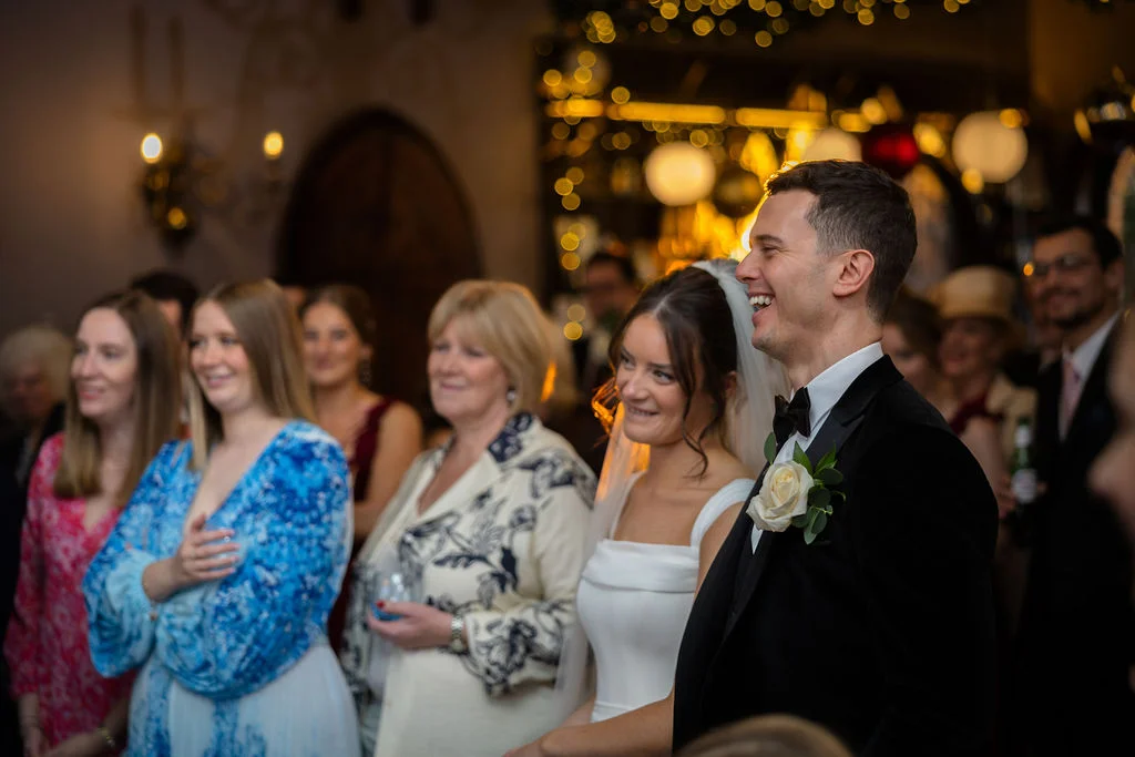 A smiling groom in a black tuxedo with a white rose buttonhole stands beside his bride while guests watch and laugh during the wedding reception.
