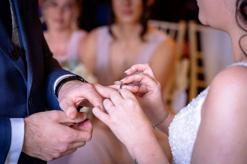 A close-up shot captures the bride gently sliding a wedding ring onto the groom's finger during the ceremony.