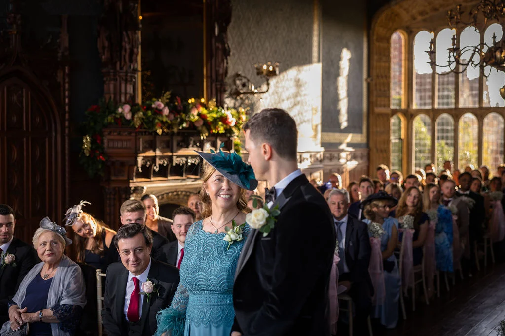 A distinguished wedding ceremony takes place inside a grand hall featuring high stained glass windows and guests seated on either side of the aisle.