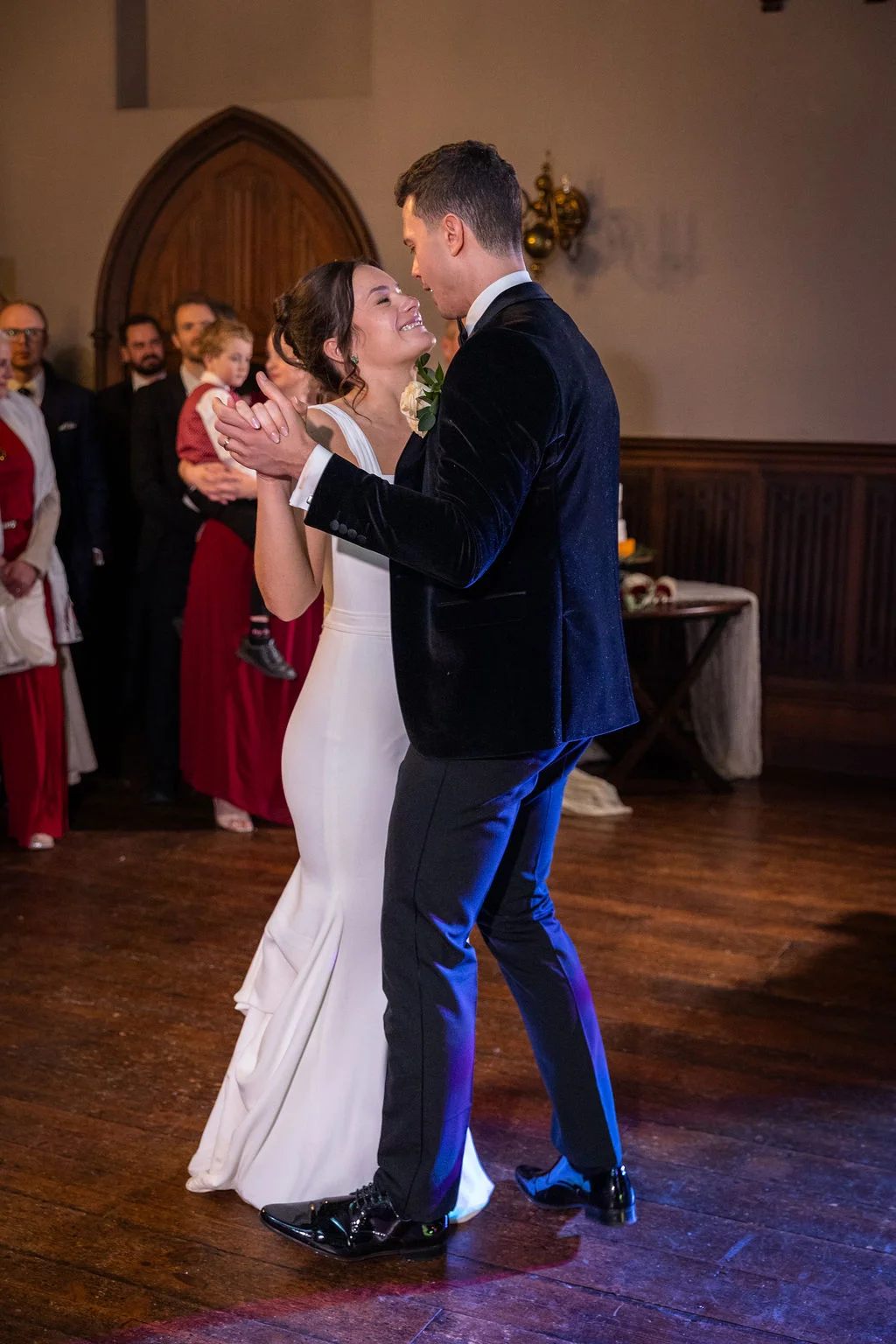Newlyweds share an intimate and joyful first dance on a polished wooden floor inside a historic hall with wooden panelling and a stone archway.