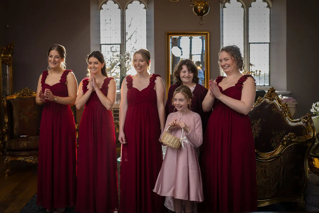 A group of bridesmaids wearing matching burgundy gowns and a young flower girl in a pink dress smile and clap inside an elegant manor house.