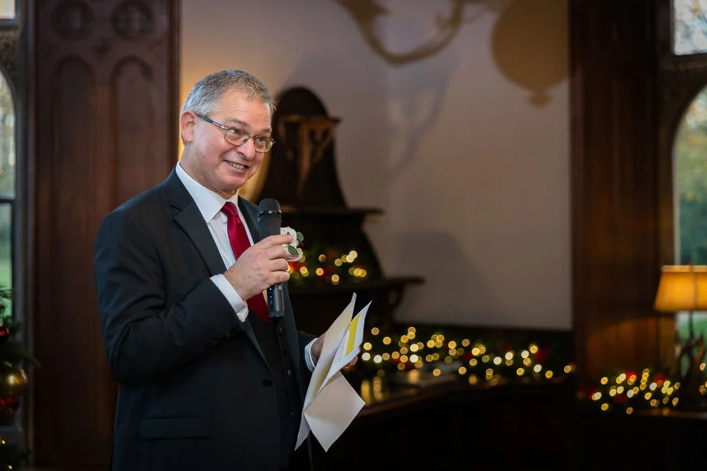 An older man wearing a dark suit and red tie smiles while holding a microphone and speech notes during wedding speeches.