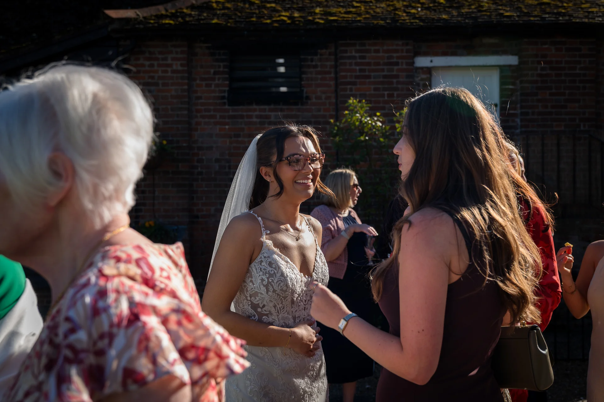A smiling bride wearing a lace wedding dress and glasses chats with guests in a sunlit outdoor courtyard with a brick building background.