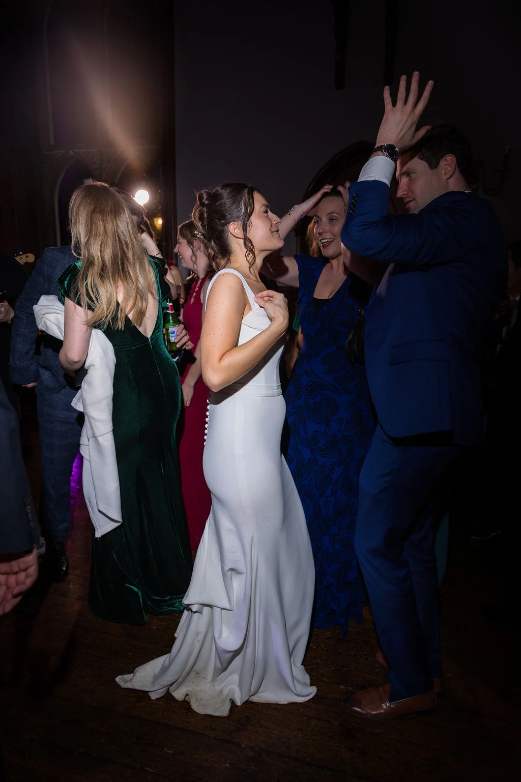 The bride in a sleek white satin gown and groom in a blue suit dance joyfully on a packed dance floor surrounded by celebrating wedding guests.