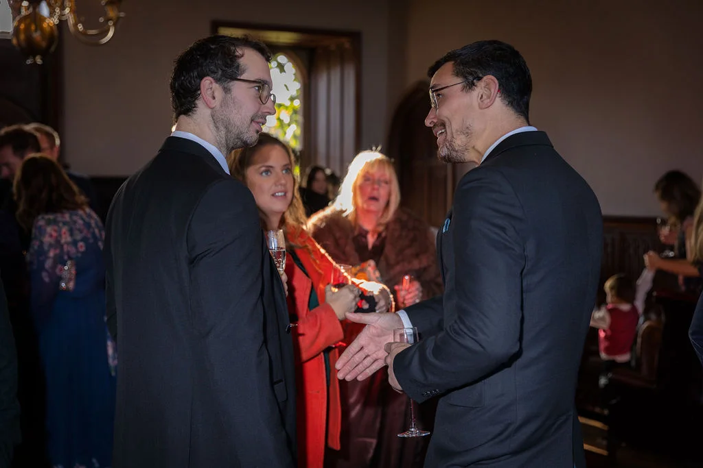 Wedding guests in smart suits and evening attire mingle and converse happily during a drinks reception inside a historic venue.