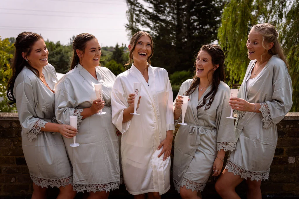 A bride in a white robe and four bridesmaids in matching sage green robes laugh together while holding champagne flutes outdoors.