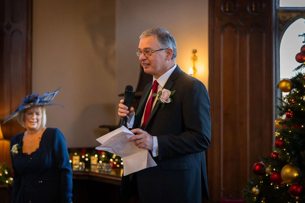 An older gentleman delivers his wedding speech holding a microphone and notes, standing beside a decorated Christmas tree while a guest watches nearby.