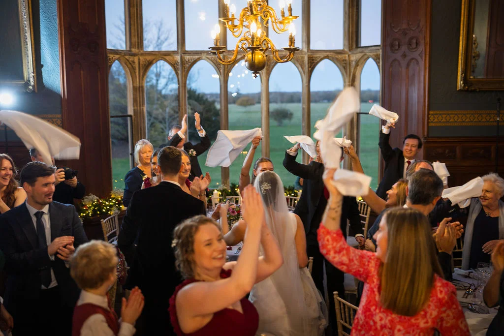 Wedding guests enthusiastically wave their white napkins in the air during the reception speeches inside a grand, wood-panelled hall with large arched windows.