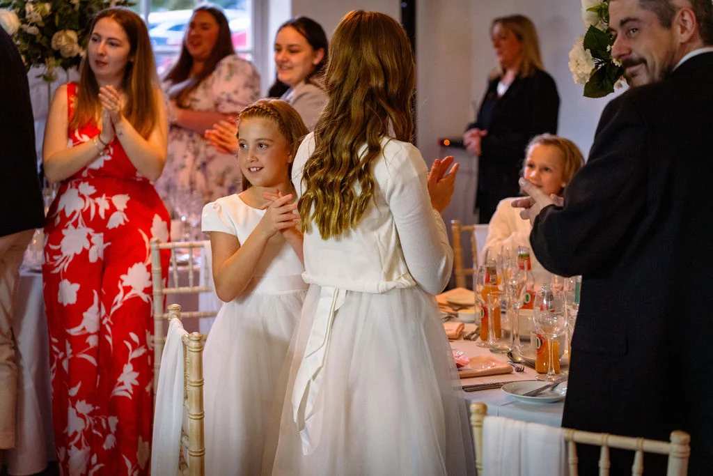 Wedding guests stand and applaud during the reception while flower girls in white dresses look on happily at the tablescape.