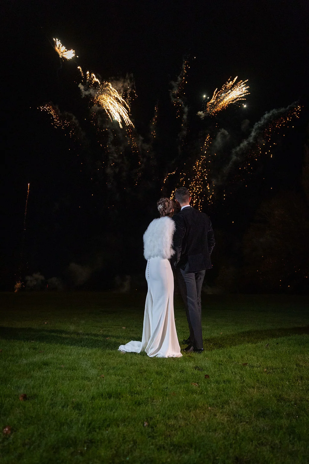 Newlyweds stand together on a grassy lawn at night, watching a vibrant fireworks display light up the dark sky.
