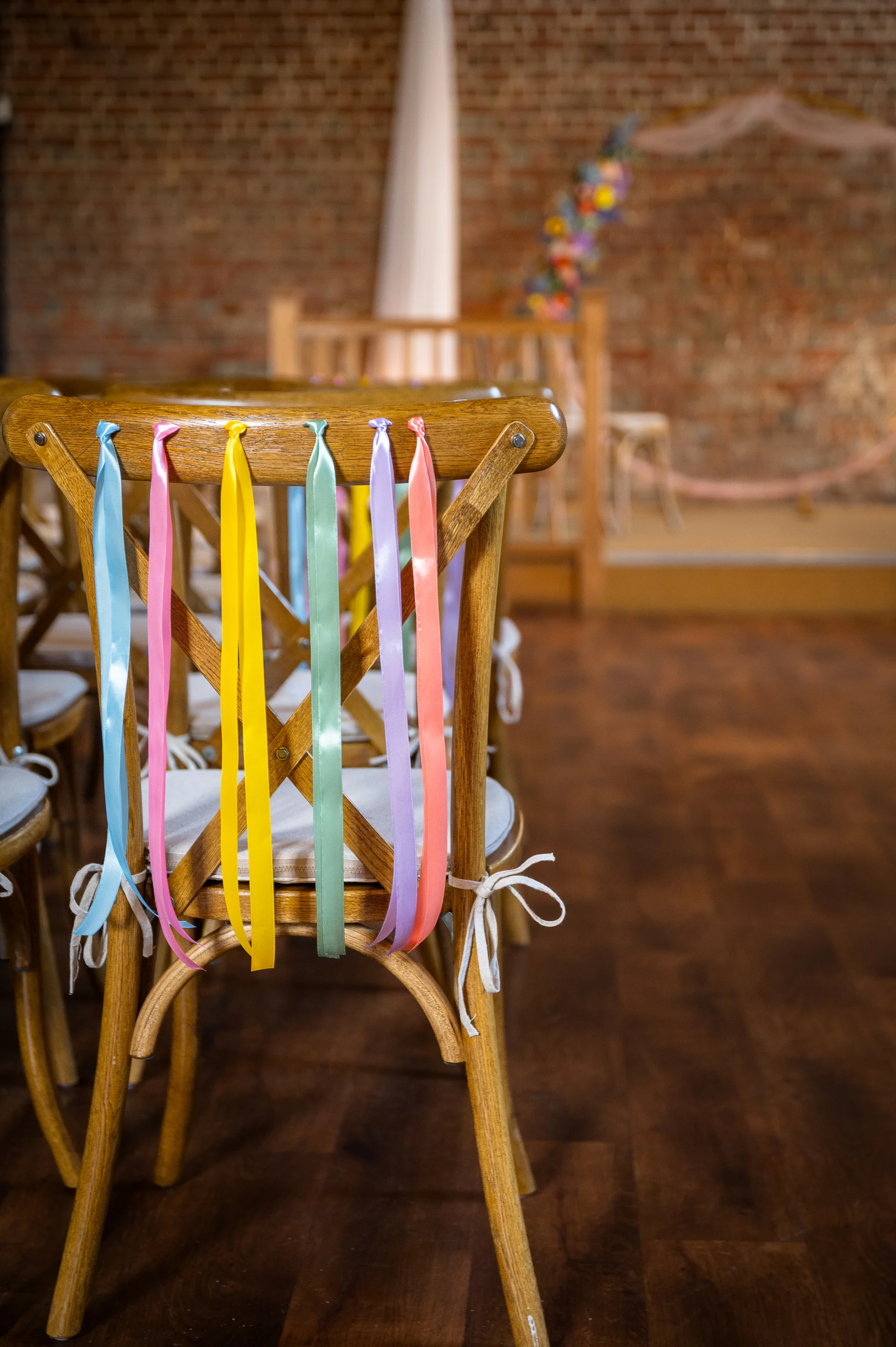 Wooden cross-back chairs decorated with colourful pastel ribbons line the aisle inside a rustic barn wedding venue.