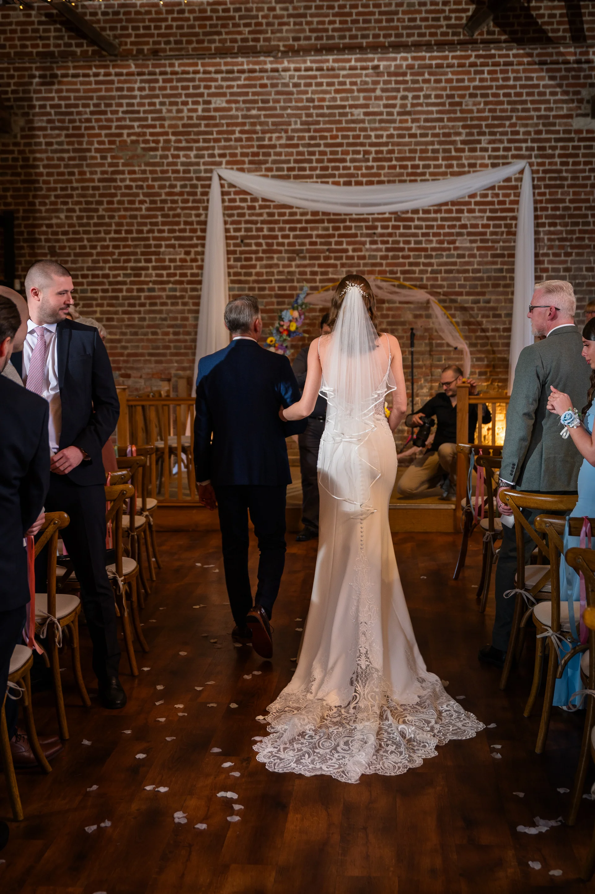 A bride walks down the aisle arm in arm with her father inside a rustic brick barn venue decorated with white drapery and scattered flower petals.
