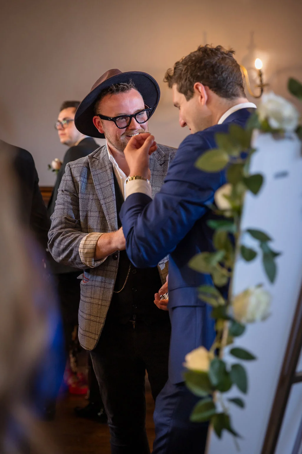 Two well-dressed grooms share a joyful and amused moment during the wedding reception while a magician performs close-up card tricks.