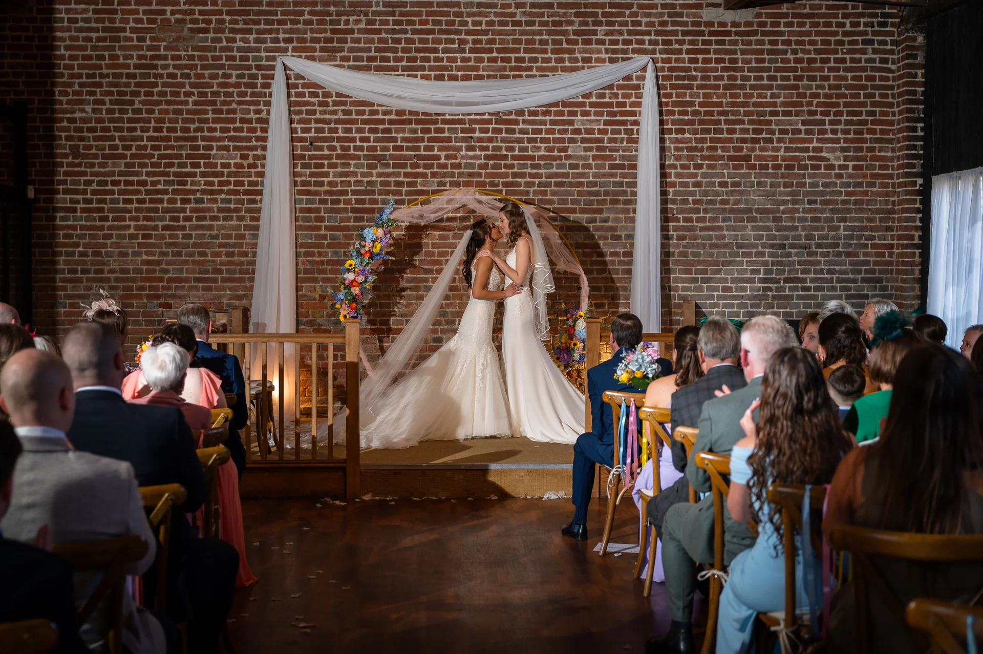 Two brides share their first kiss as a married couple beneath a circular floral arch inside a rustic brick barn, watched by seated guests.