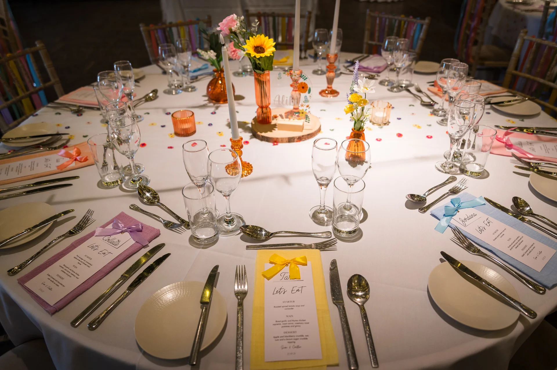 A beautifully set round wedding breakfast table features colourful menus, vibrant floral arrangements with sunflowers, taper candles, and delicate glassware.
