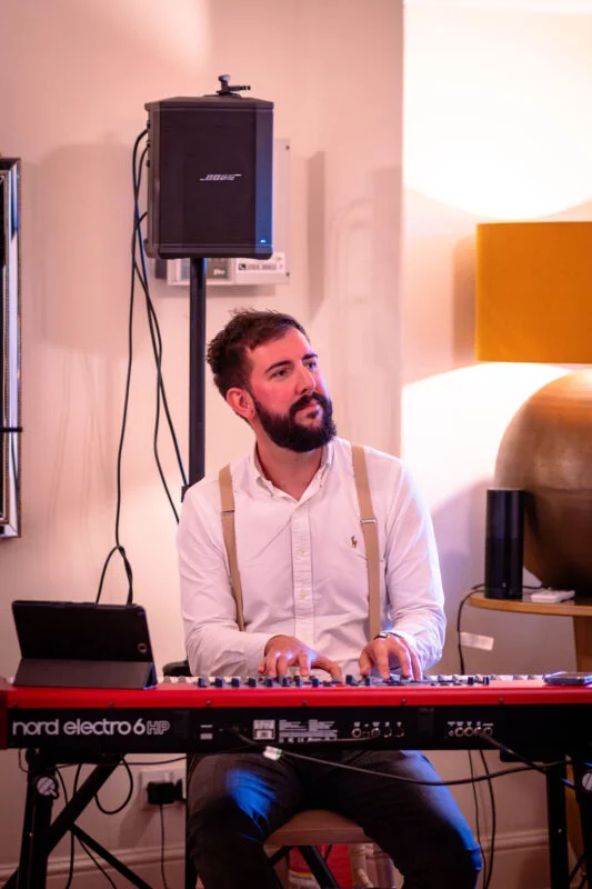A bearded musician wearing a white shirt and braces sits indoors playing a red stage keyboard during a wedding celebration.