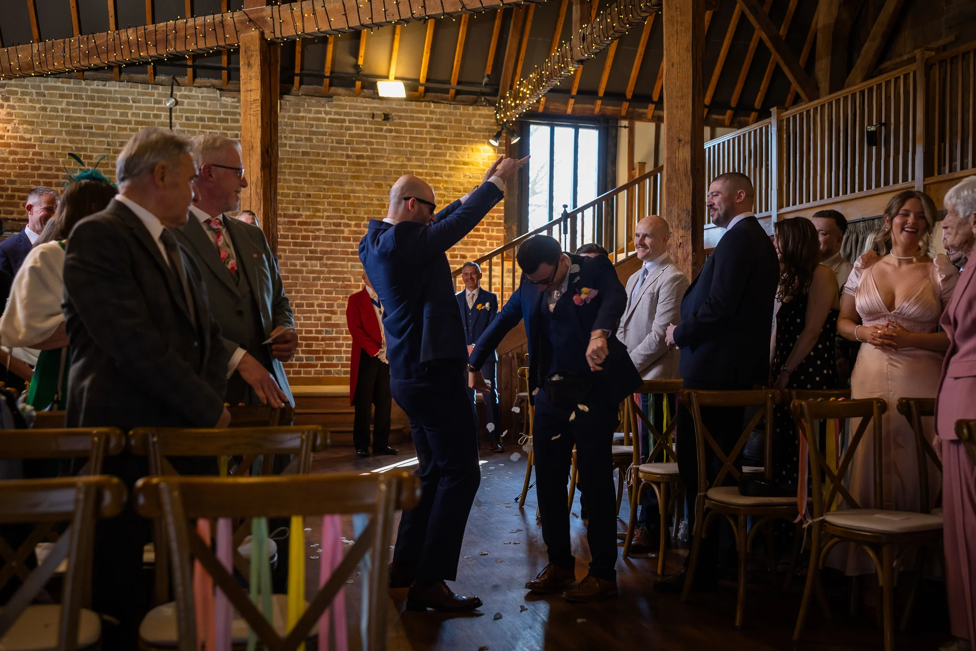 Two grooms in navy suits celebrate amidst laughing wedding guests inside a rustic oak framed barn adorned with fairy lights.