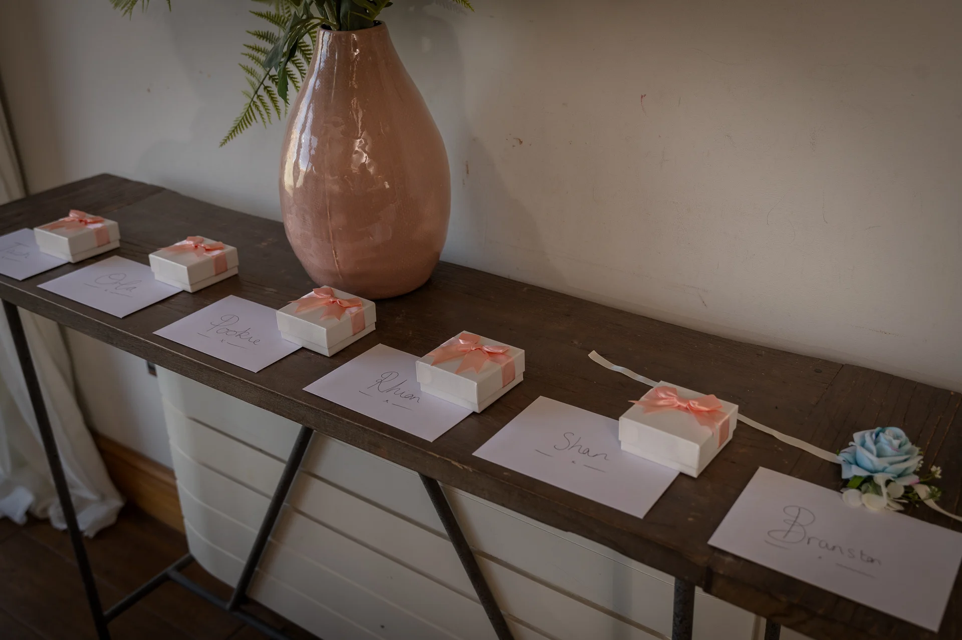 A rustic wooden console table displays a row of white gift boxes tied with peach ribbons alongside handwritten place cards and a large pink ceramic vase.