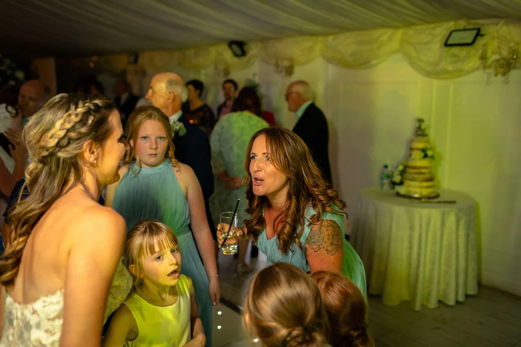 A candid shot capturing a joyful wedding guest speaking animatedly with the bride inside a marquee decorated with draped ceiling fabric and a wedding cake table in the background.