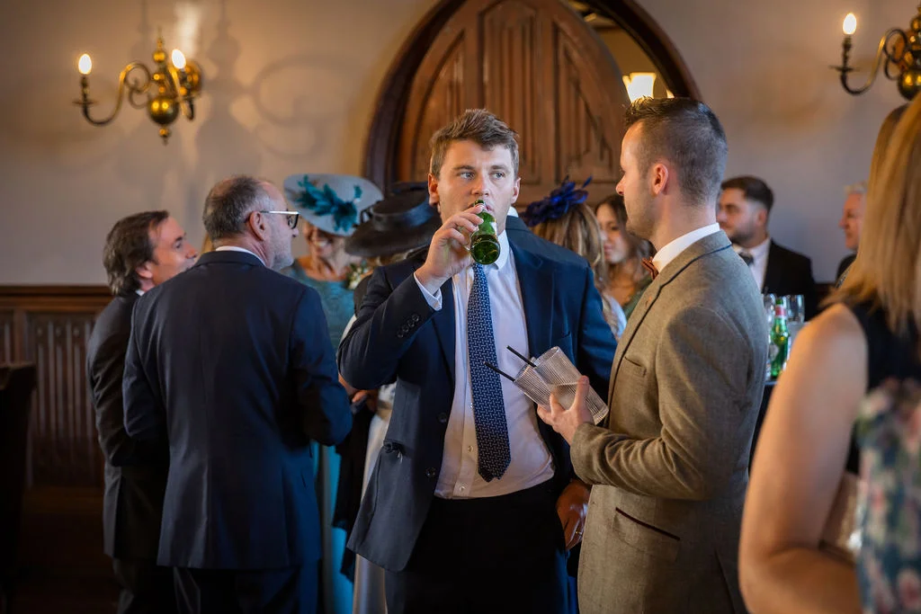 Wedding guests mingling and enjoying drinks in a stately country house reception room with wood panelled walls and a stone archway.