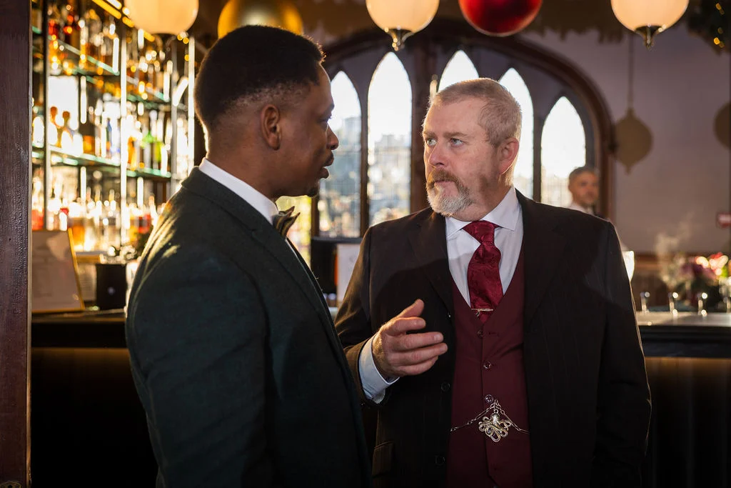 Two well-dressed men converse earnestly near a backlit bar during a wedding reception, one wearing a dark jacket and the other a three-piece suit with a burgundy waistcoat and tie.