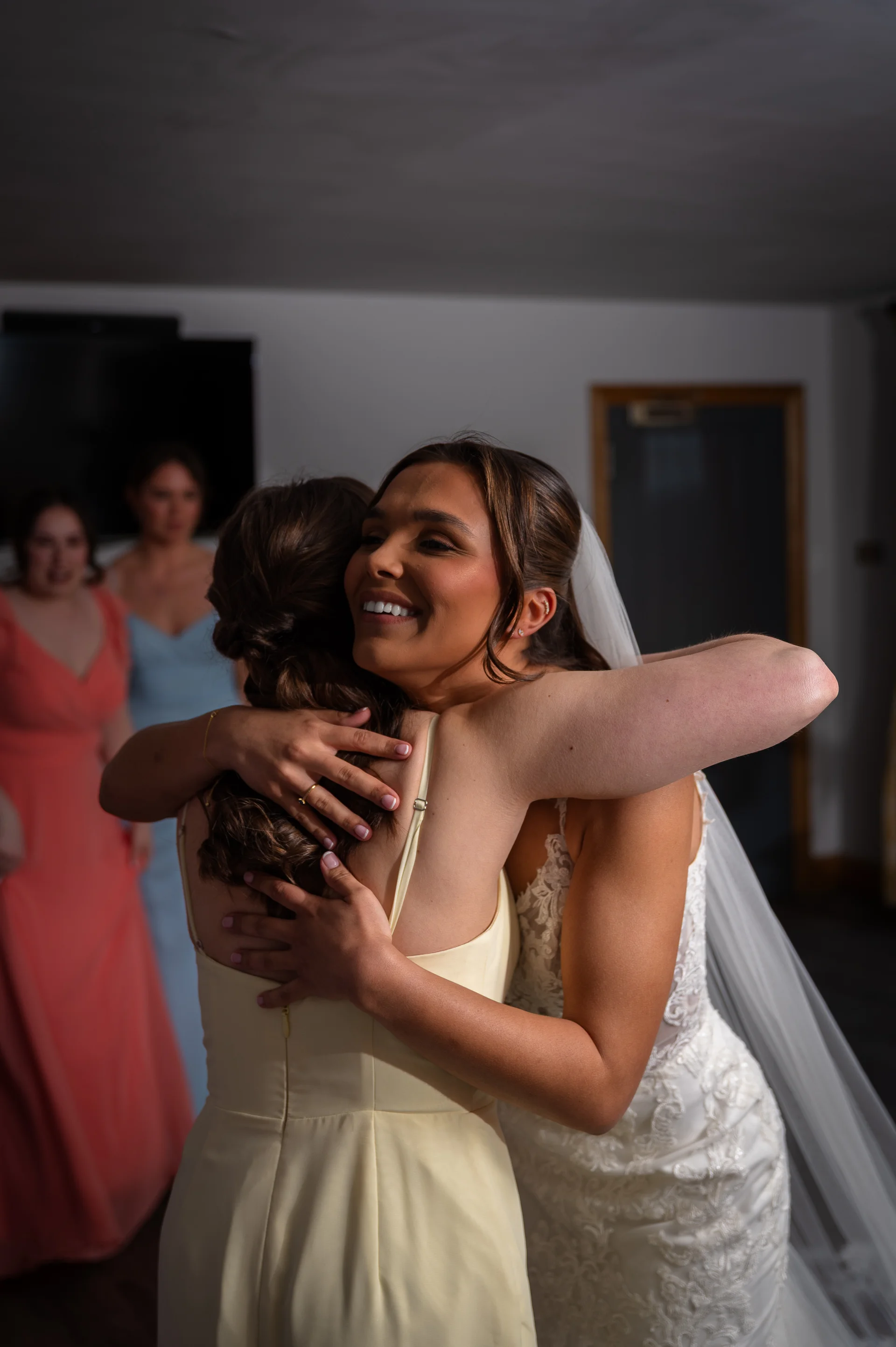 A glowing bride in a lace gown and veil embraces a bridesmaid wearing a yellow dress during bridal preparations while other bridesmaids look on.