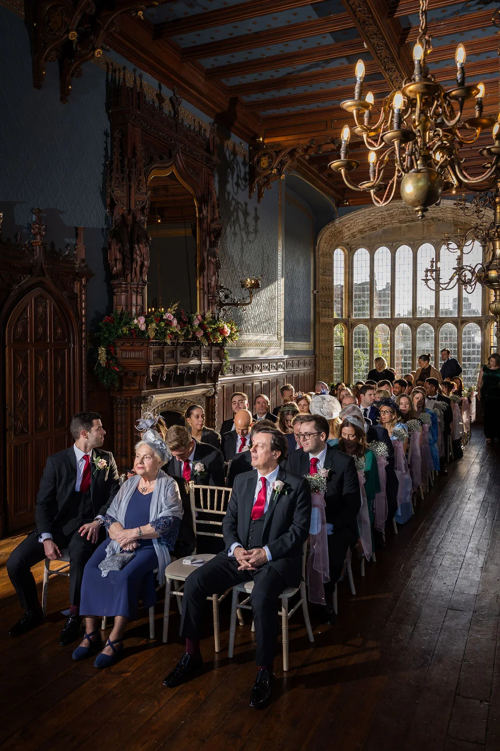 Guests are seated in a grand stately home with wooden panelling and large arched windows for a traditional wedding ceremony.
