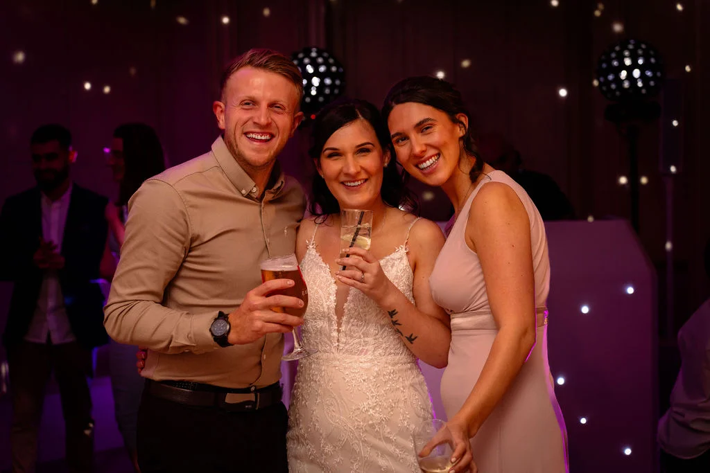 A smiling bride, groom, and guest pose together holding drinks during the evening reception under atmospheric venue lighting.