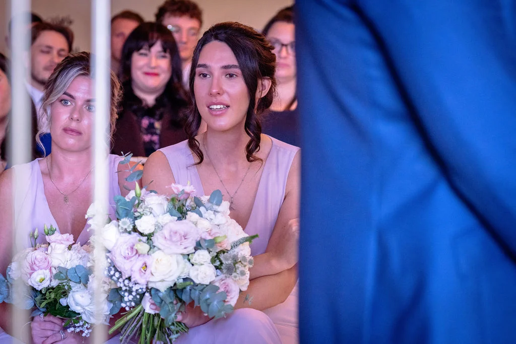 A bridesmaid in a lilac dress holding a hand-tied bouquet of roses and eucalyptus looks intently towards the front during a wedding ceremony.