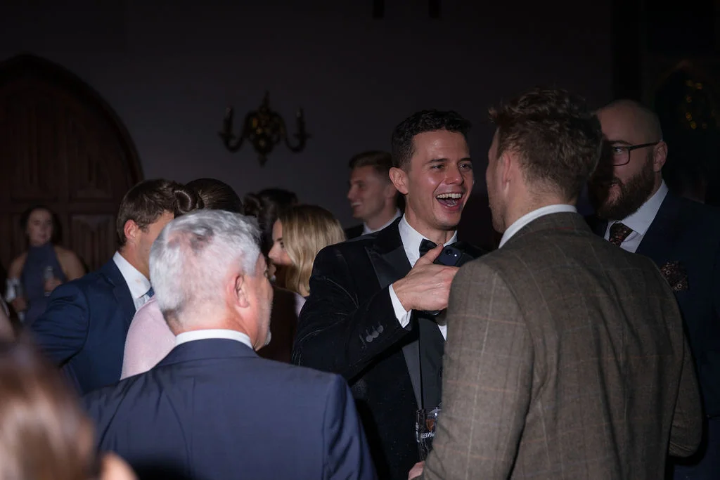 A smiling groom in a black tuxedo laughs heartily while mingling with guests during the evening wedding reception.