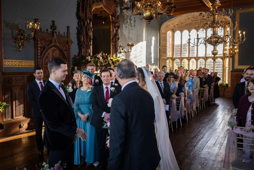 The groom smiles at the bride as she approaches the altar during a traditional ceremony inside an ornate, wood-panelled historic hall.