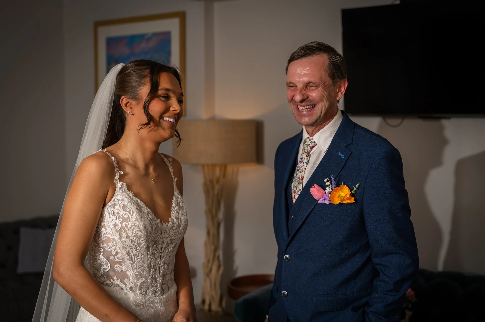 A radiant bride in a lace wedding dress smiling warmly as she shares a joyful first look moment with her father wearing a blue suit.