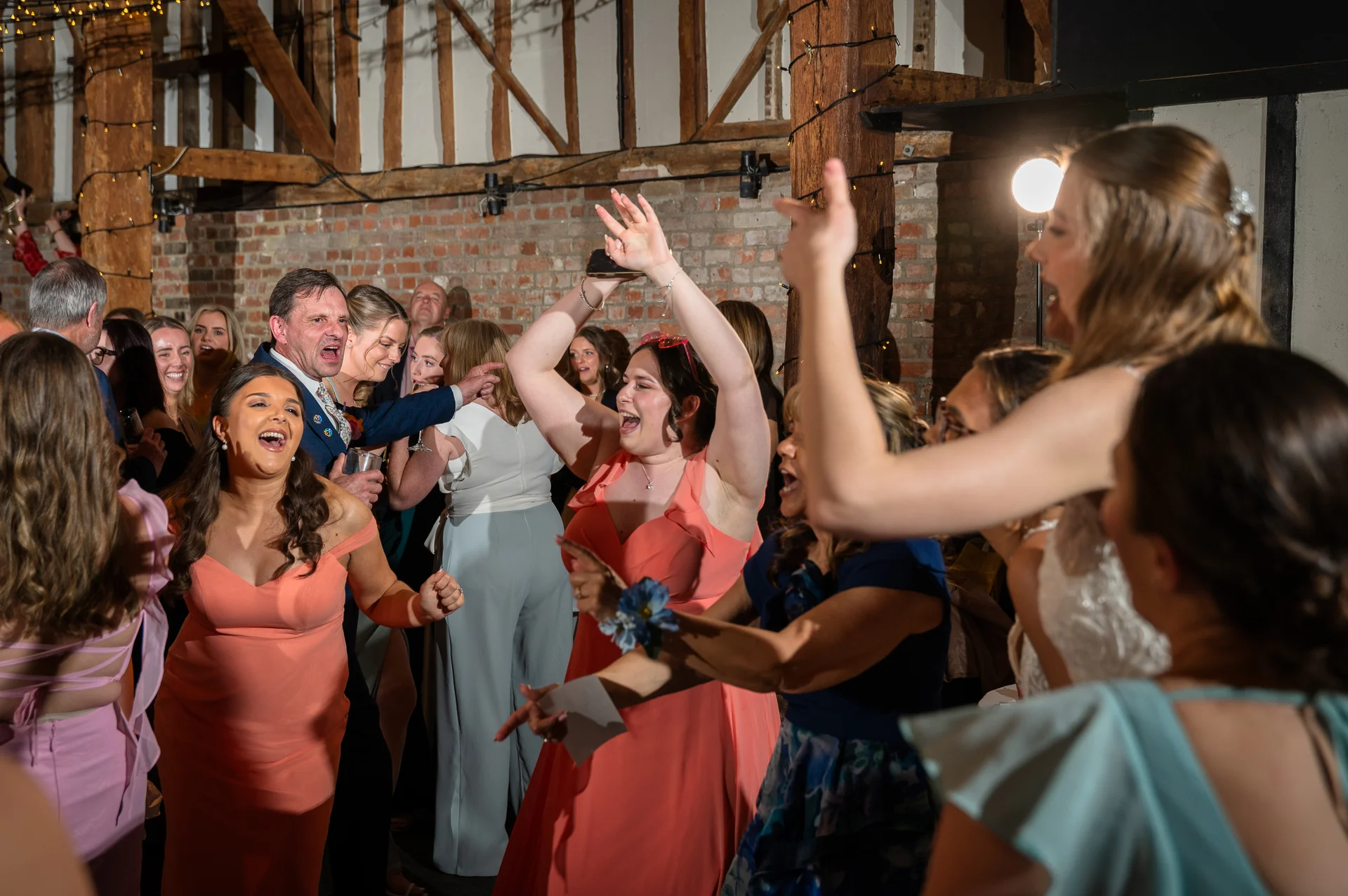 A lively group of wedding guests celebrating on a crowded dance floor inside a rustic brick barn adorned with fairy lights.