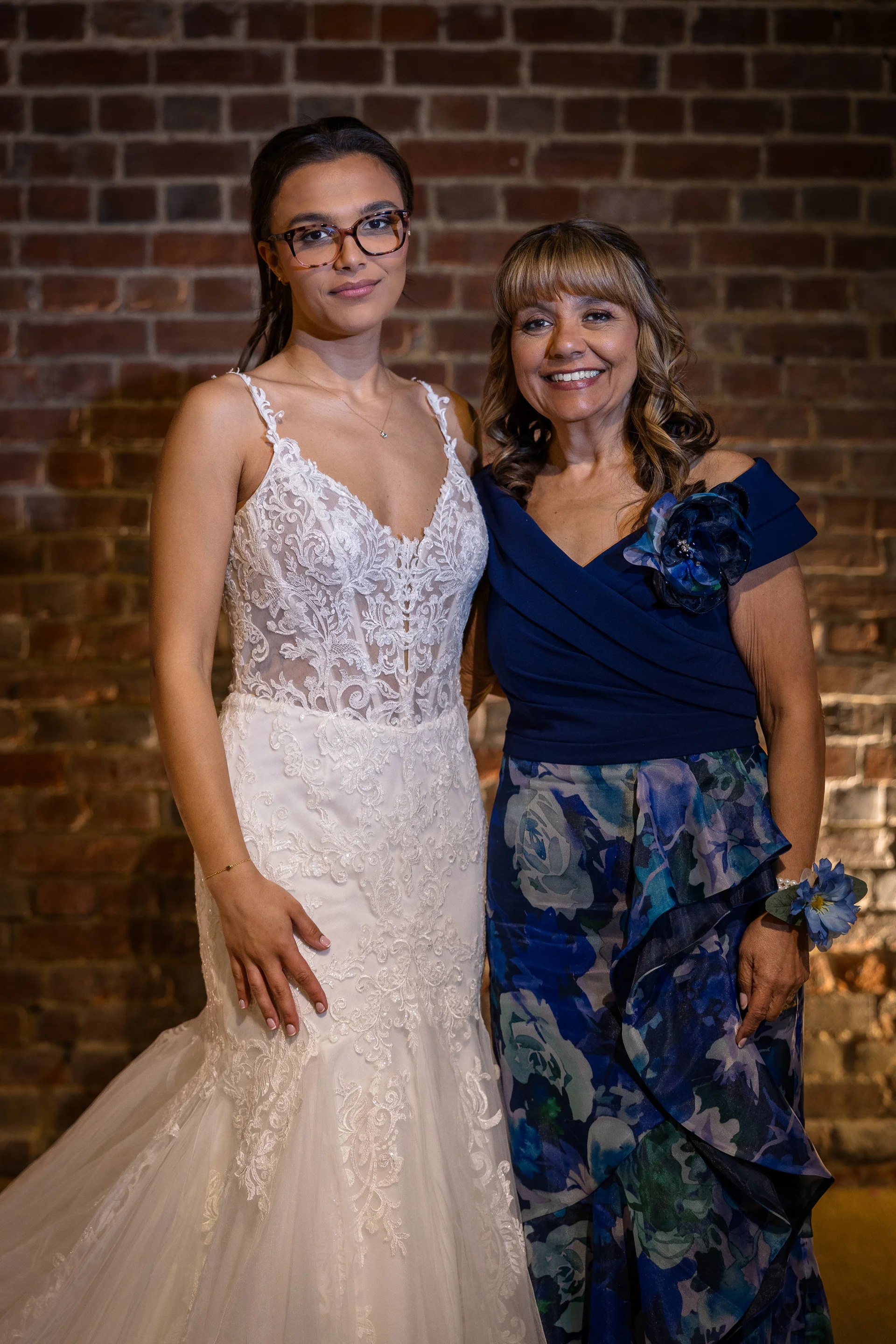 A smiling bride wearing glasses and a lace mermaid wedding dress stands alongside her mother in a blue patterned dress in front of an exposed brick wall.