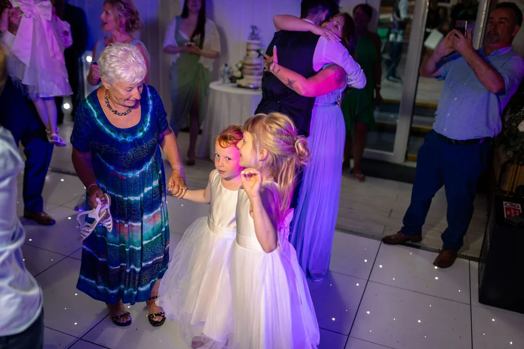 Wedding guests and flower girls dance joyfully on an illuminated dance floor under atmospheric blue lighting while others embrace in the background.