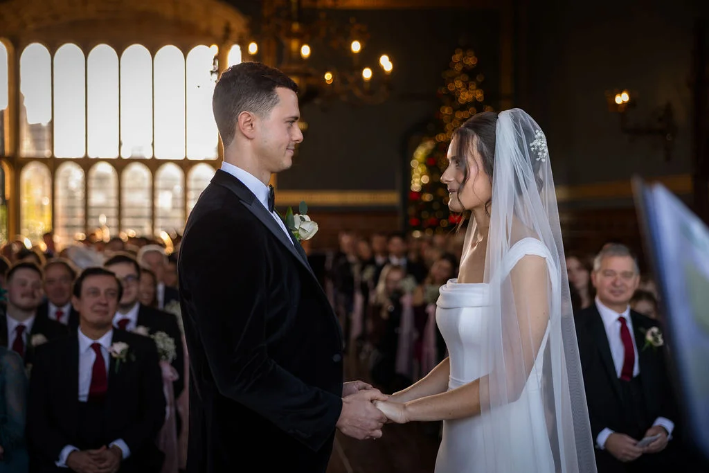 The bride and groom hold hands and gaze into each other's eyes during their indoor winter wedding ceremony inside a grand historic venue.