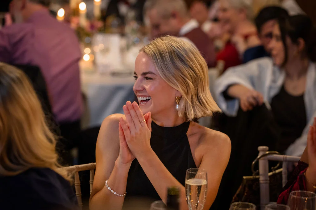 A blonde wedding guest with a chic bob hairstyle laughs joyfully and claps her hands during the wedding reception speeches.
