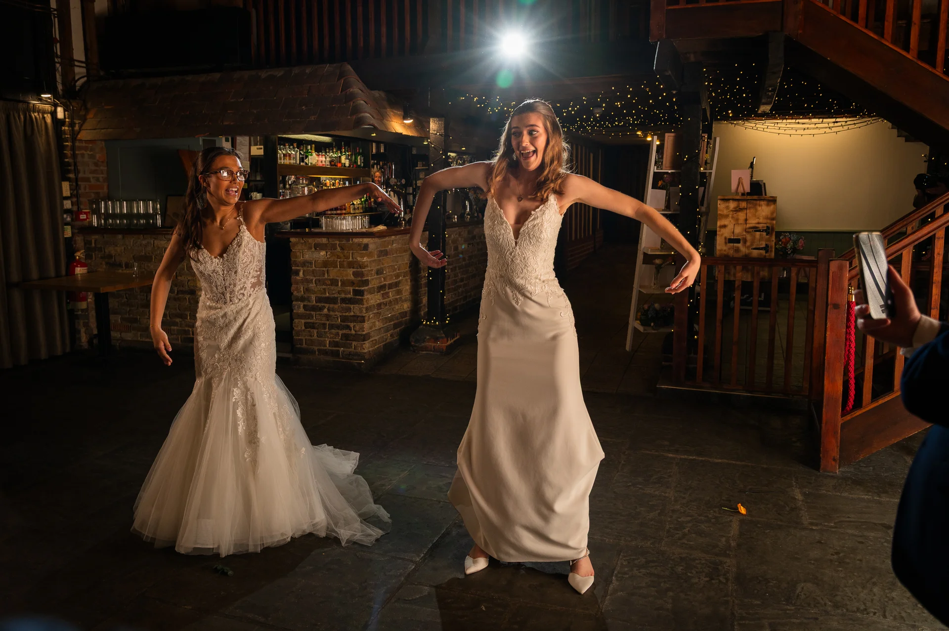 Two smiling brides in wedding dresses joyfully dance in a rustic barn venue illuminated by overhead string lights.