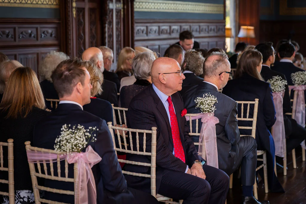 Guests are seated in a wood-panelled room with chairs decorated with pink bows and gypsophila flowers during a wedding ceremony.