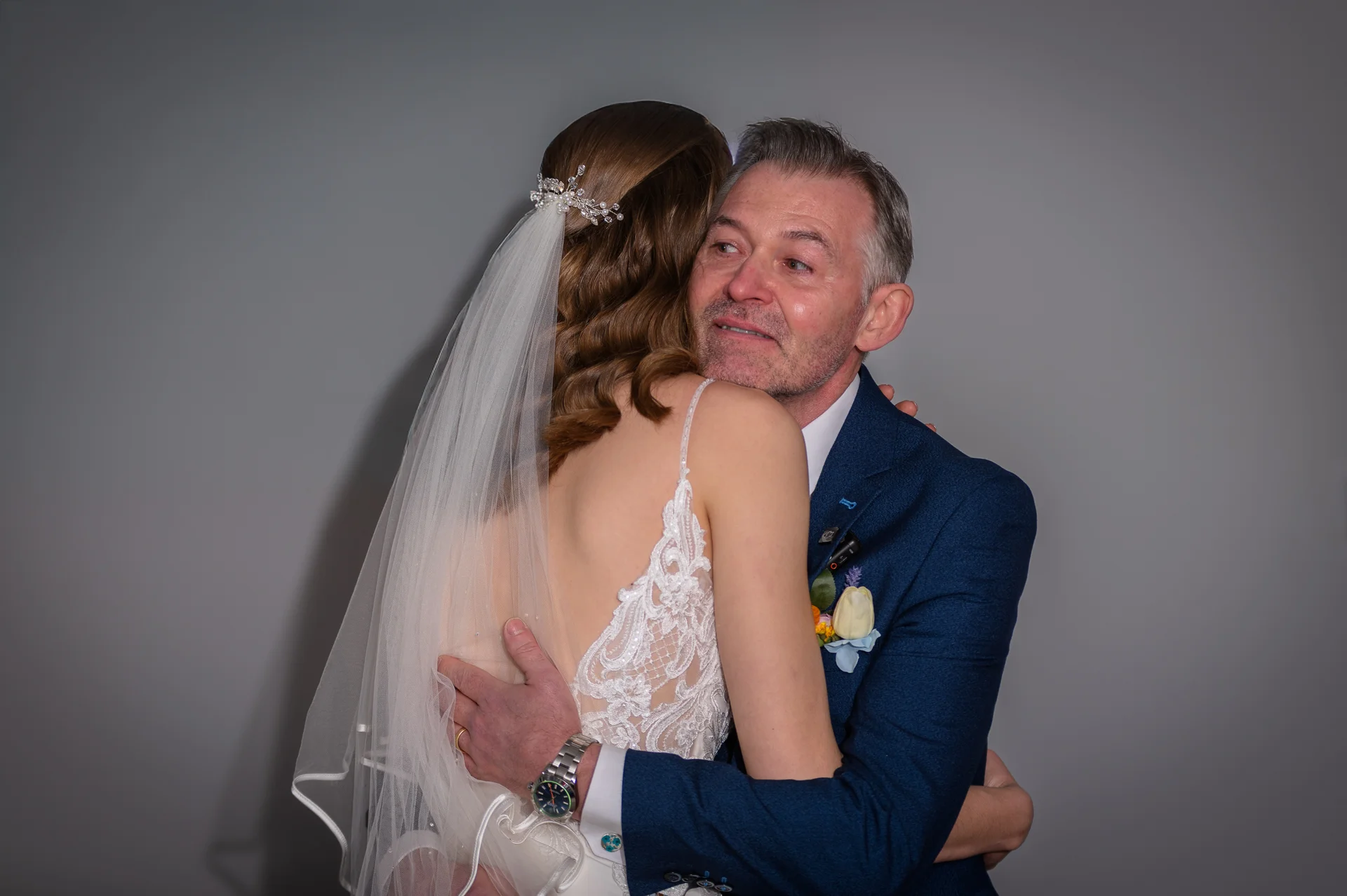 An emotional father of the bride shares a tender, tearful hug with his daughter while wearing a blue suit and boutonniere.