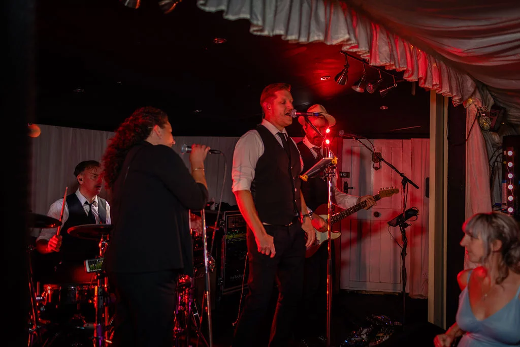 A live wedding band performs on a stage inside a marquee under dramatic red lighting, with a guest watching from the foreground.