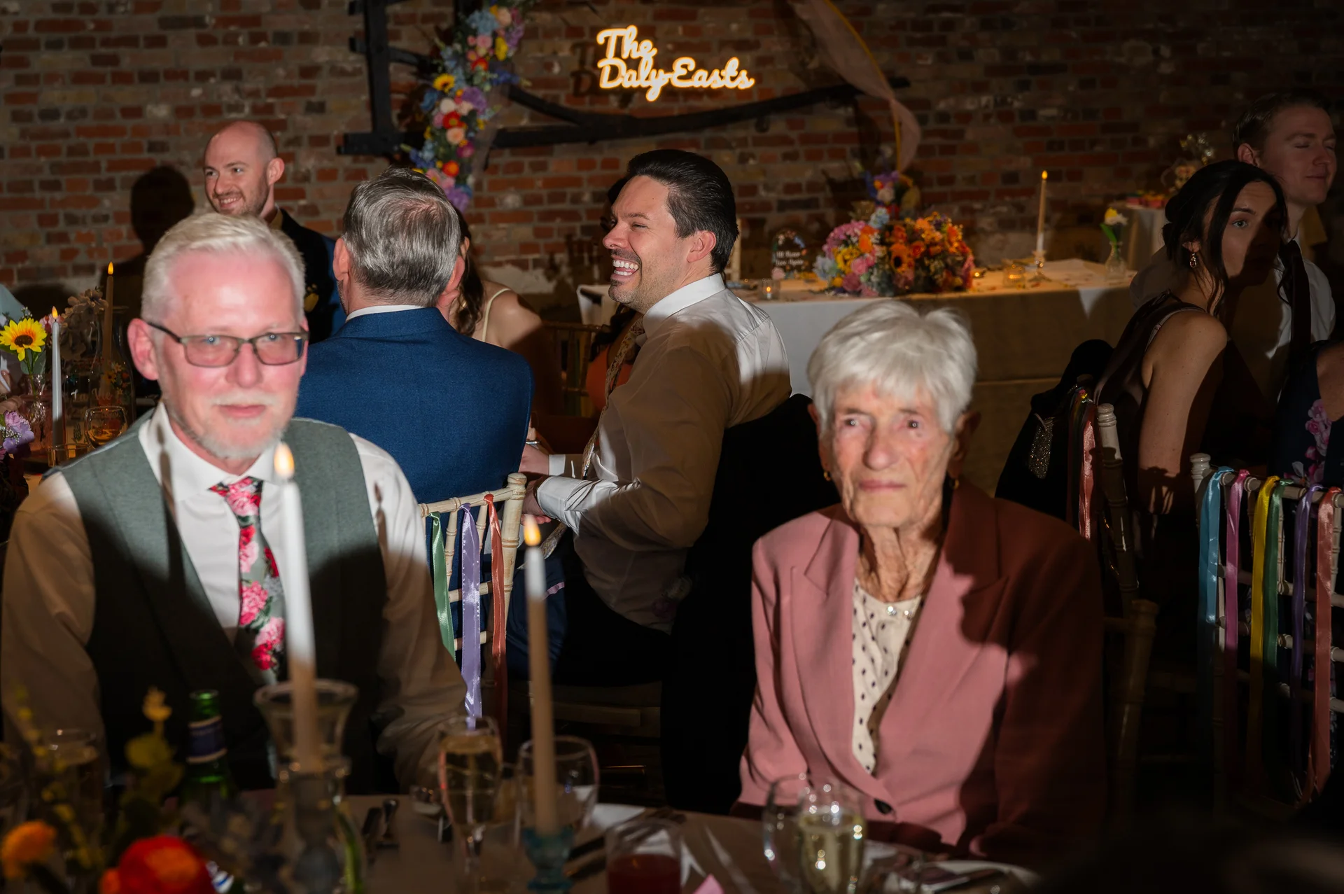 A candid evening wedding reception shot featuring guests seated at dining tables laughing and enjoying speeches in front of a rustic exposed brick wall.