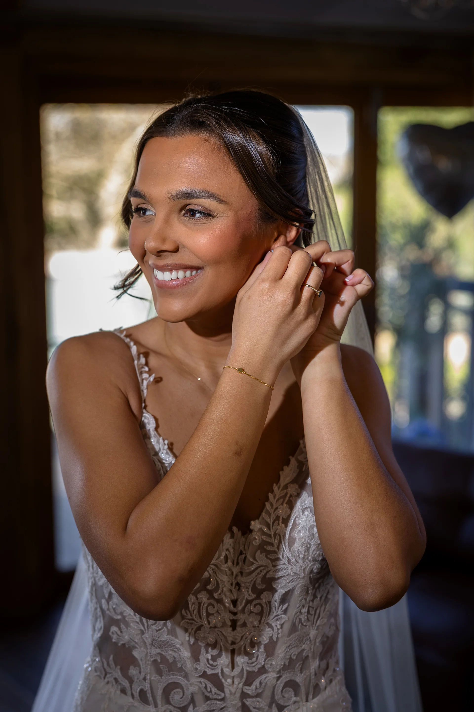 A smiling bride puts on her earrings while getting ready on her wedding morning, wearing an intricate lace dress with a plunging neckline.