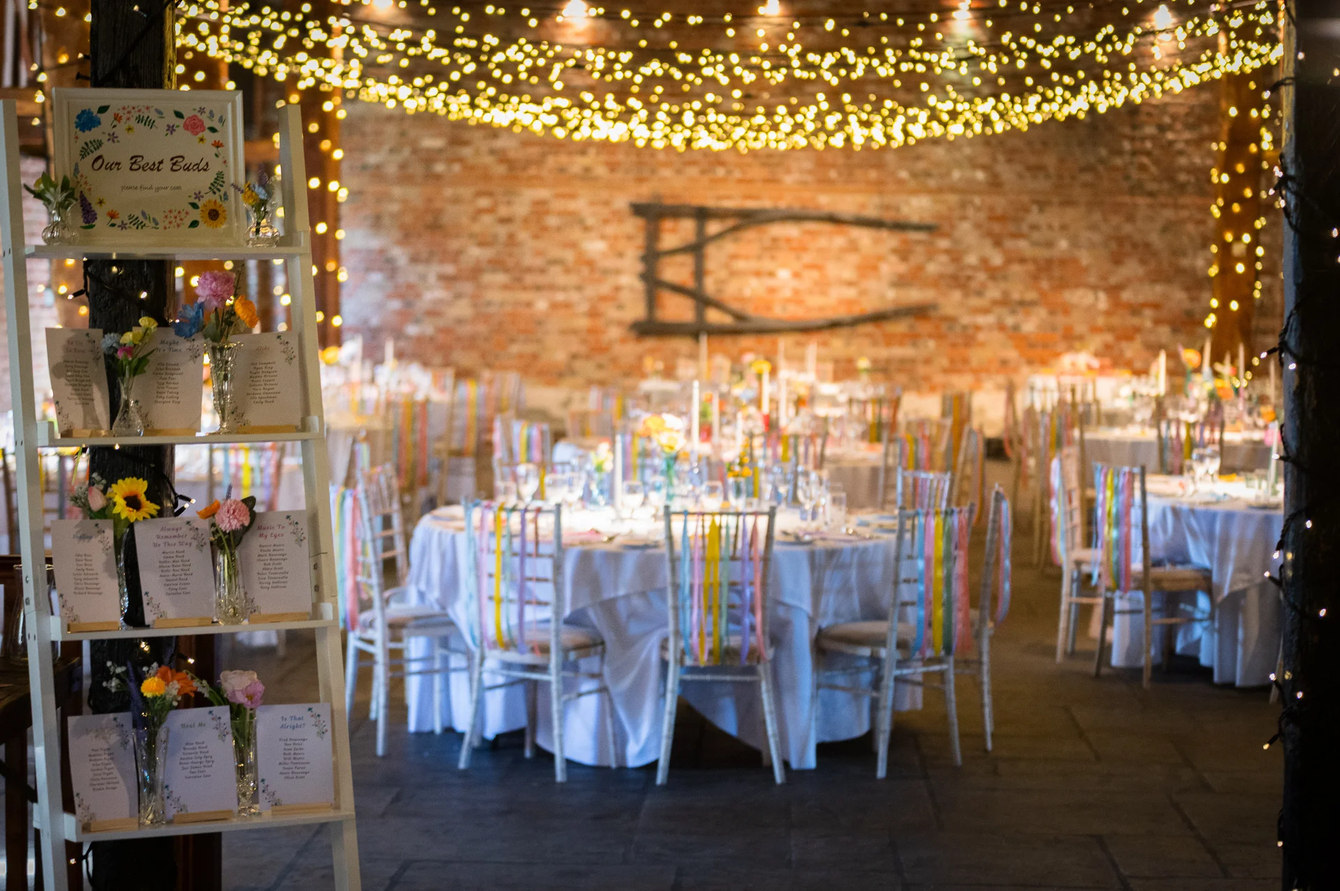 A rustic brick barn wedding reception venue features round tables decorated with hanging ribbons on chairs and a glowing canopy of fairy lights overhead.