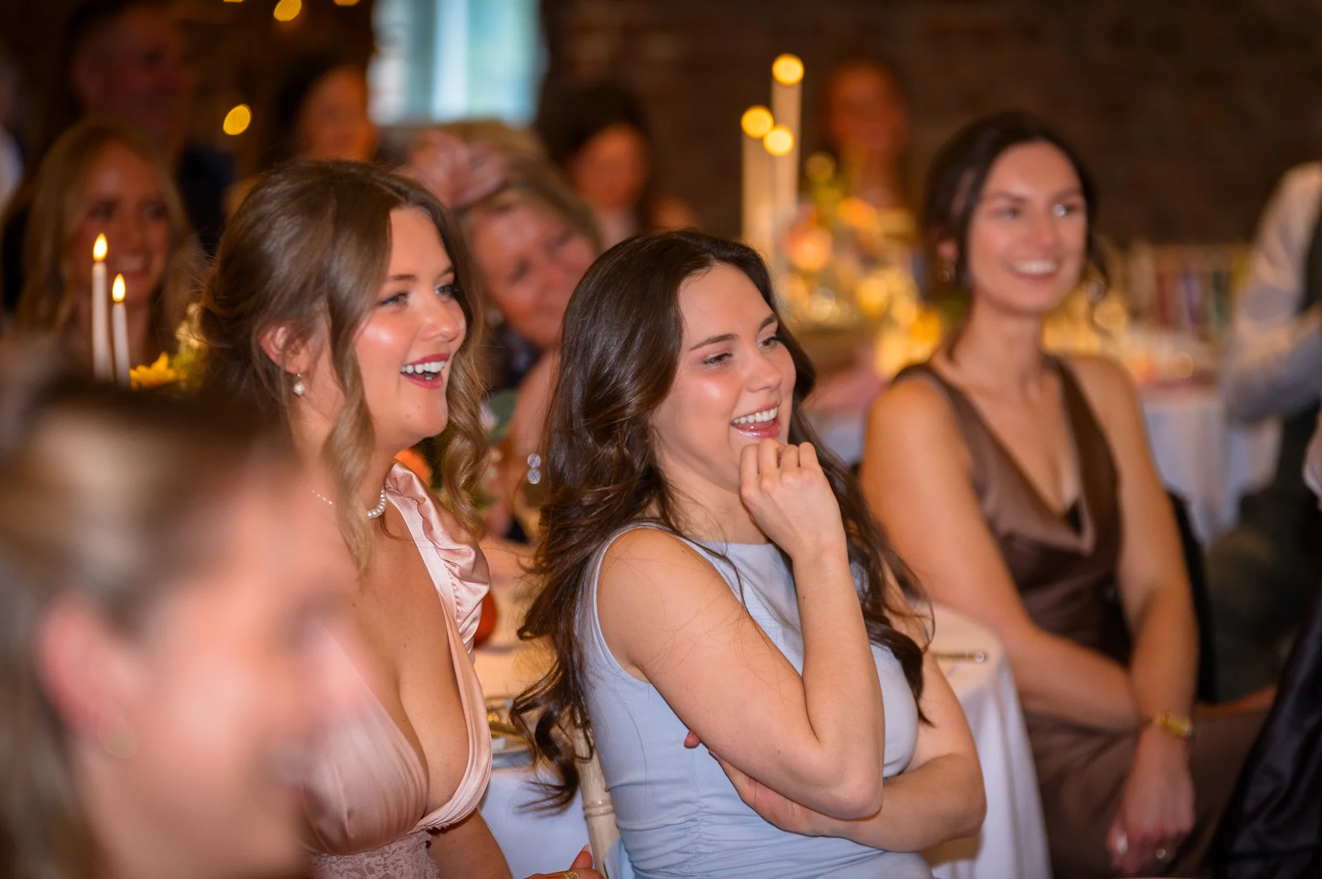 Several wedding guests are seated at a candlelit reception table, laughing and enjoying the speeches with warm ambient lighting.