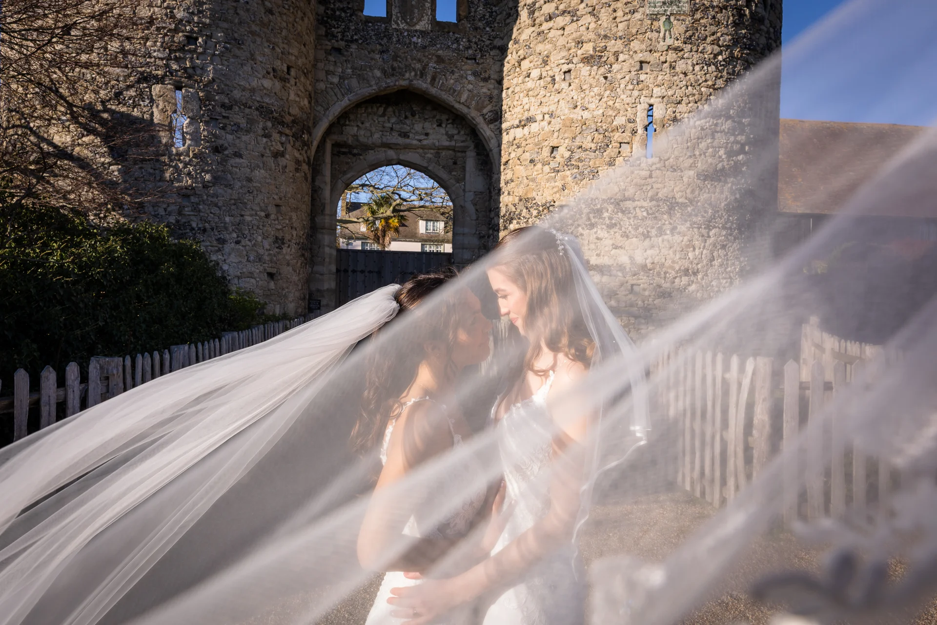 Two brides embrace tenderly outdoors in front of a historic stone castle gate, framed dramatically by a flowing sheer veil billowing across the foreground.