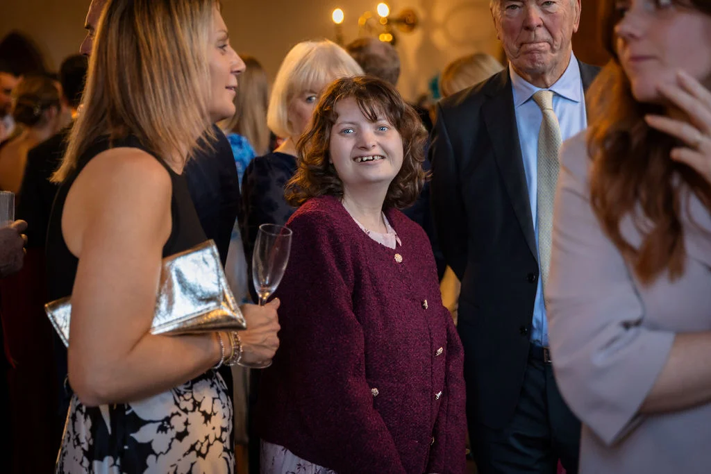 Candid photograph of wedding guests mingling at a drinks reception, featuring a smiling woman holding a champagne flute while wearing a textured burgundy jacket.