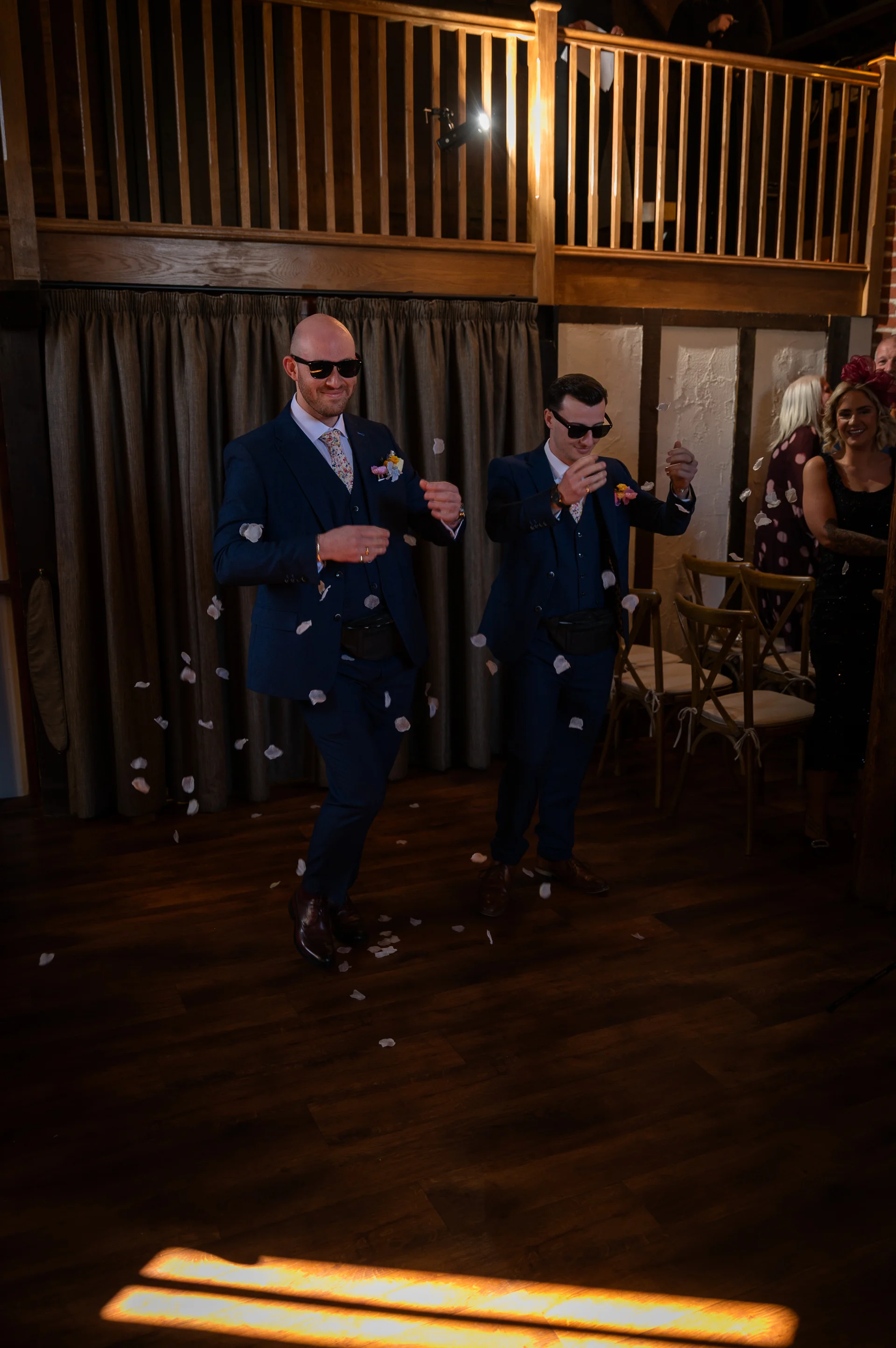 Two grooms in matching navy suits and sunglasses dance joyfully as white confetti and petals fall around them inside a rustic barn venue.