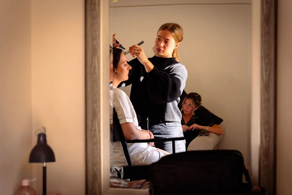 A bride sits in a makeup chair while a professional makeup artist applies cosmetics, all captured through a large mirror reflection during morning bridal preparations.